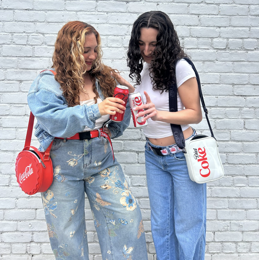 Two young women wearing Coca-Cola and Diet Coke seatbelt belts, holding Coke cans, styled with a Coca-Cola bottle-cap crossbody bag and Diet Coke crossbody bag against a white brick wall.